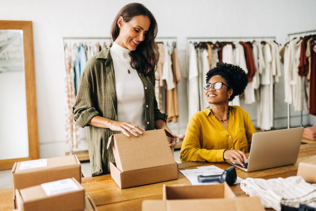 Duas empreendedoras, uma sorrindo enquanto organiza caixas de produtos e a outra trabalhando em um laptop, gerenciam um negócio de varejo, exemplificando o empreendedorismo feminino e o e-commerce.