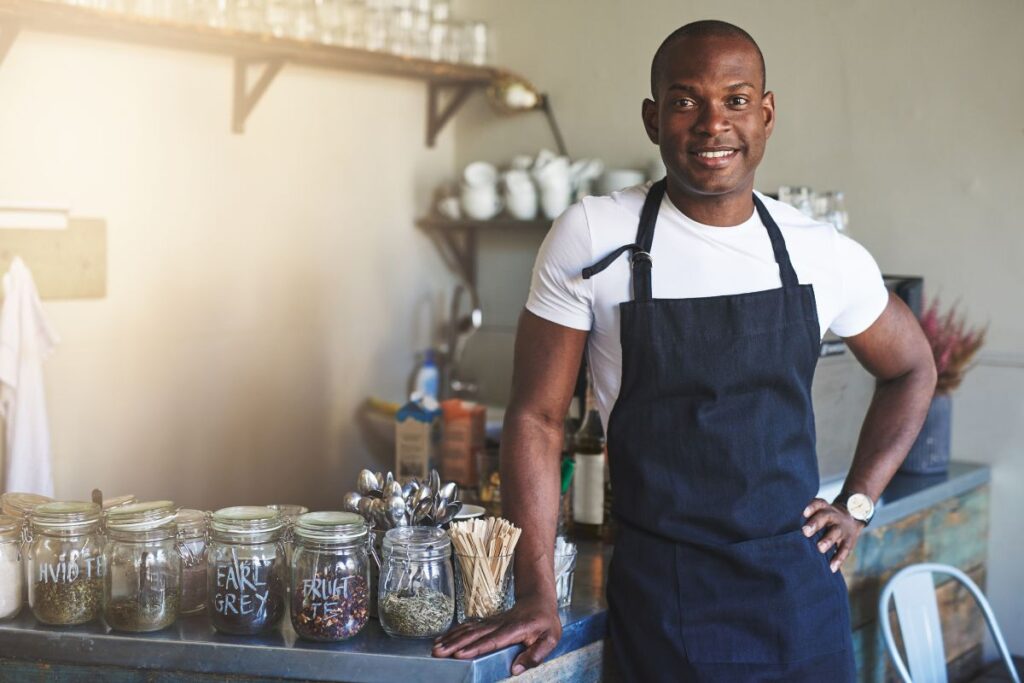 Um empreendedor negro sorridente, vestindo um avental, posa com confiança atrás do balcão de seu café ou loja de chás, com potes de ingredientes ao fundo.