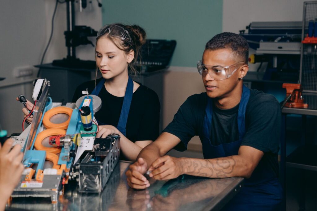 Dois jovens aprendizes, um homem e uma mulher, trabalhando em um projeto de robótica ou engenharia, usando óculos de proteção.