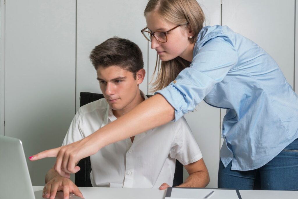 Jovem aprendiz sendo orientada por uma colega ou supervisora em frente a um laptop.