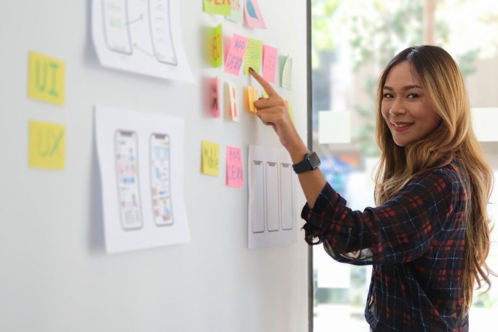 Mulher sorridente interagindo com um quadro branco cheio de notas adesivas e esboços de interfaces de aplicativos, representando o planejamento de um projeto.