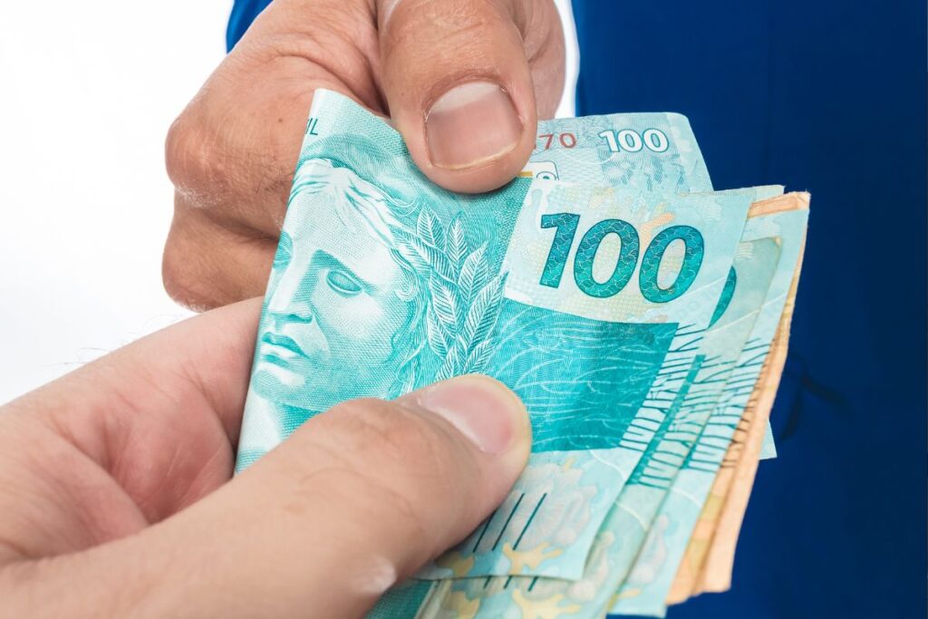 Uma mão entregando notas de cem reais para outra mão, simbolizando o pagamento do 13º salário.