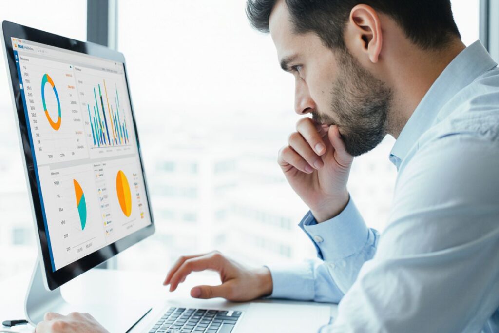 Um homem com barba, vestindo uma camisa social azul clara, olhando pensativamente para um monitor de computador que exibe gráficos e dados de negócios.