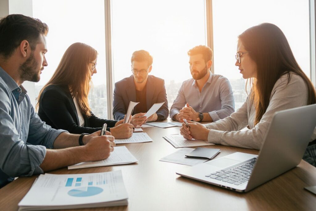 Cinco profissionais em uma reunião de negócios, discutindo estratégias e analisando documentos, com a luz do sol entrando por grandes janelas ao fundo.