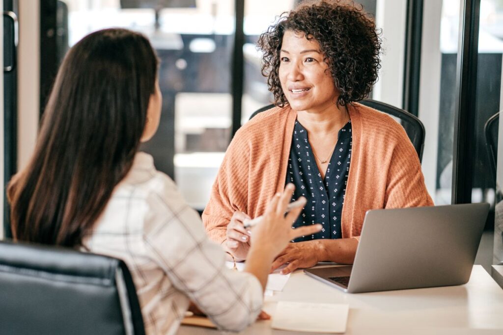 Duas mulheres em um diálogo engajado durante uma entrevista de emprego, com um laptop na mesa.