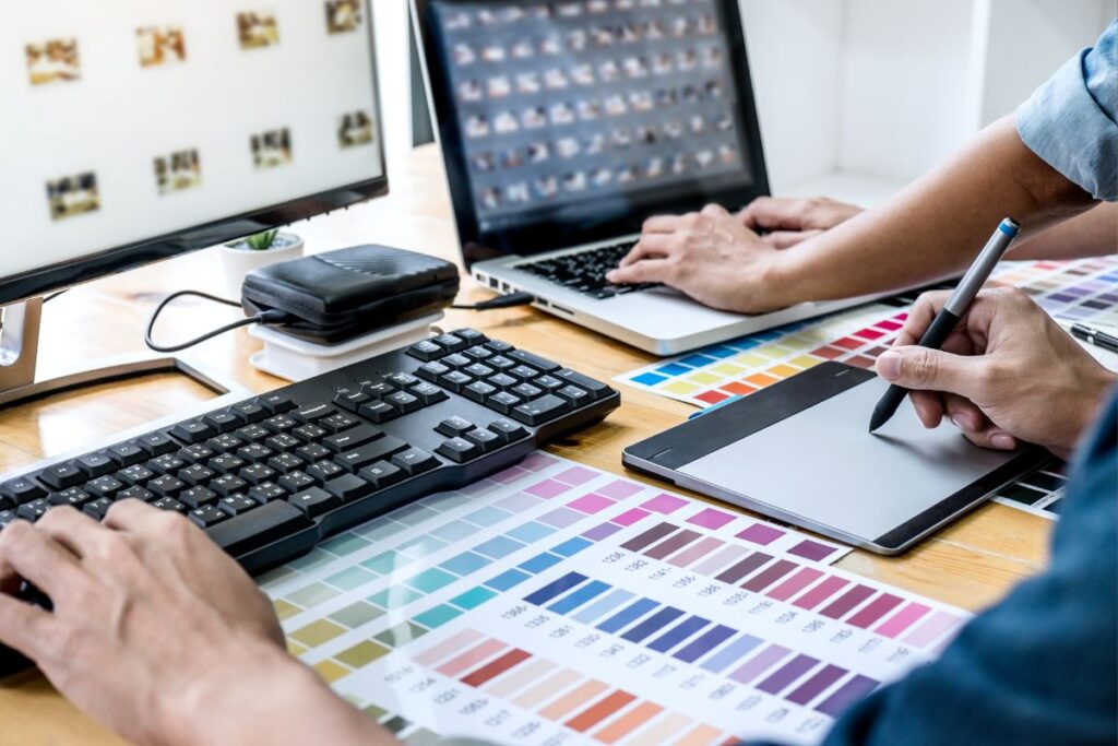 Duas pessoas trabalhando em conjunto em um escritório doméstico, usando computadores e um tablet gráfico com amostras de cores, ilustrando a colaboração no design gráfico em home office.
