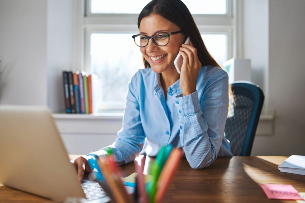 Mulher em chamada de celular sorrindo e digitando no laptop.