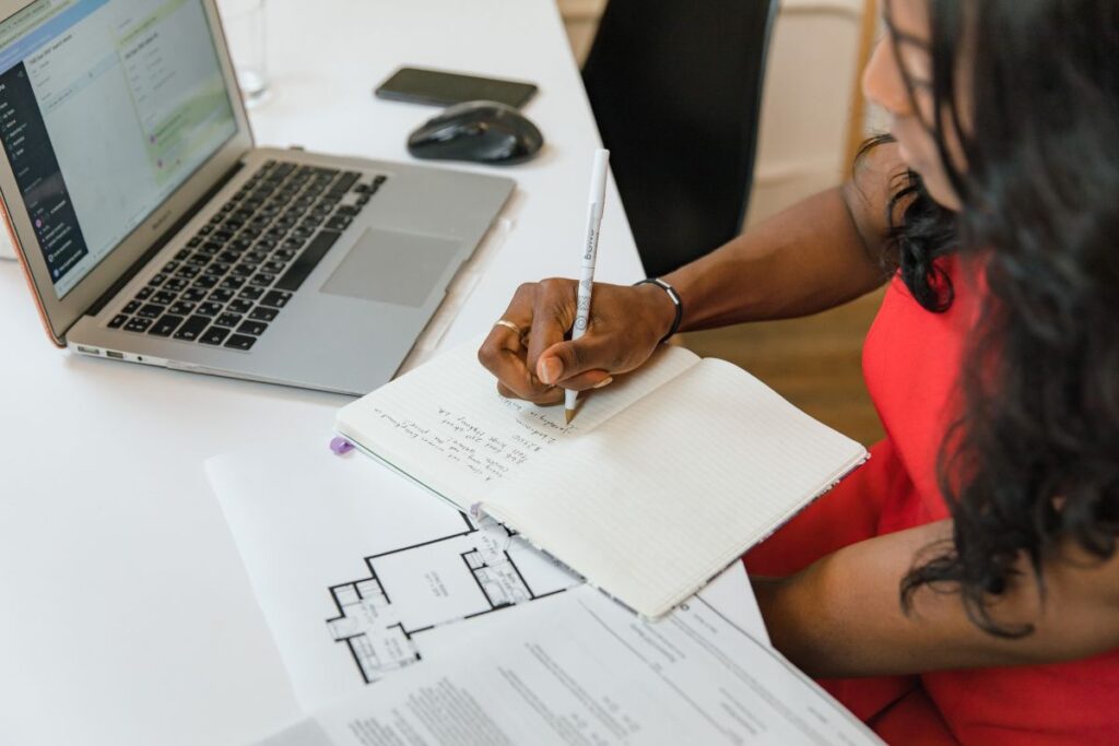 Mulher negra com cabelo preso, vestindo blusa vermelha, sentada à mesa com um laptop e cadernos, escrevendo em um deles com uma caneta branca. Ela está focada em suas anotações, com plantas baixas visíveis em outras folhas na mesa.