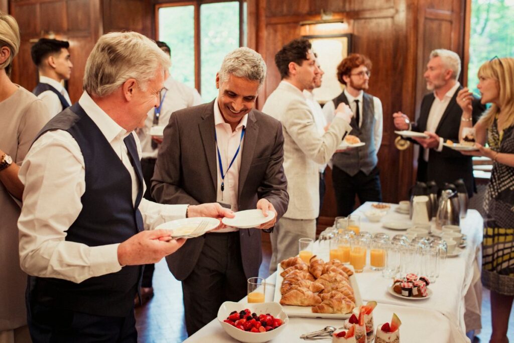 Grupo de profissionais interagindo e desfrutando de um coffee break durante um evento, com foco em dois homens sorrindo enquanto pegam comida.