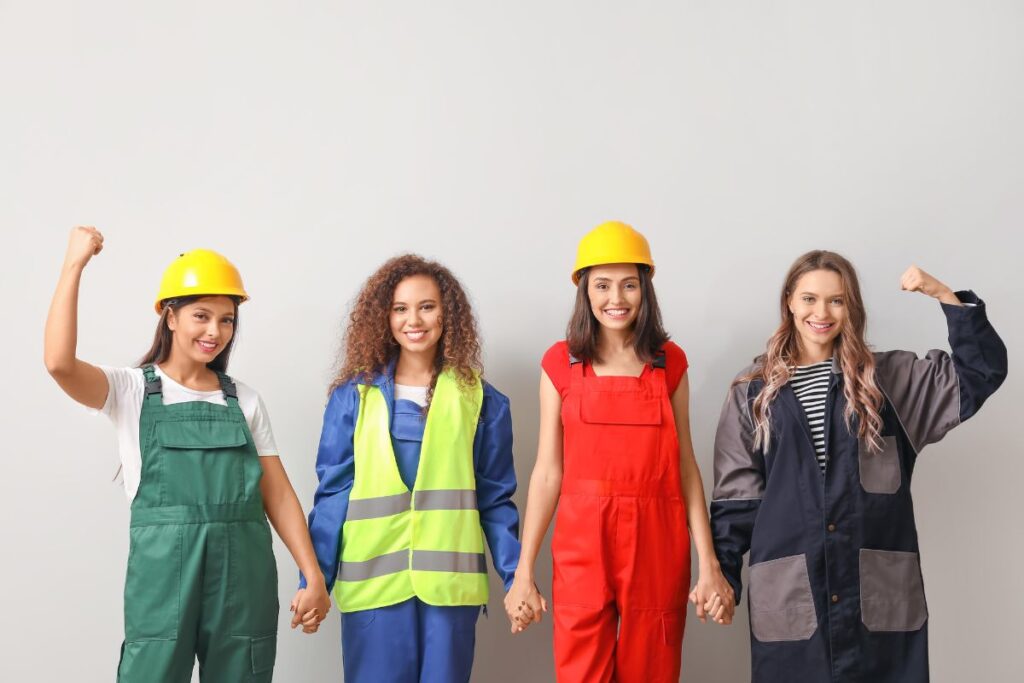 Quatro mulheres trabalhadoras de diferentes etnias, vestindo uniformes de trabalho e capacetes de segurança, de mãos dadas e sorrindo, algumas com punhos erguidos, simbolizando união e empoderamento feminino no trabalho e o direito ao descanso.