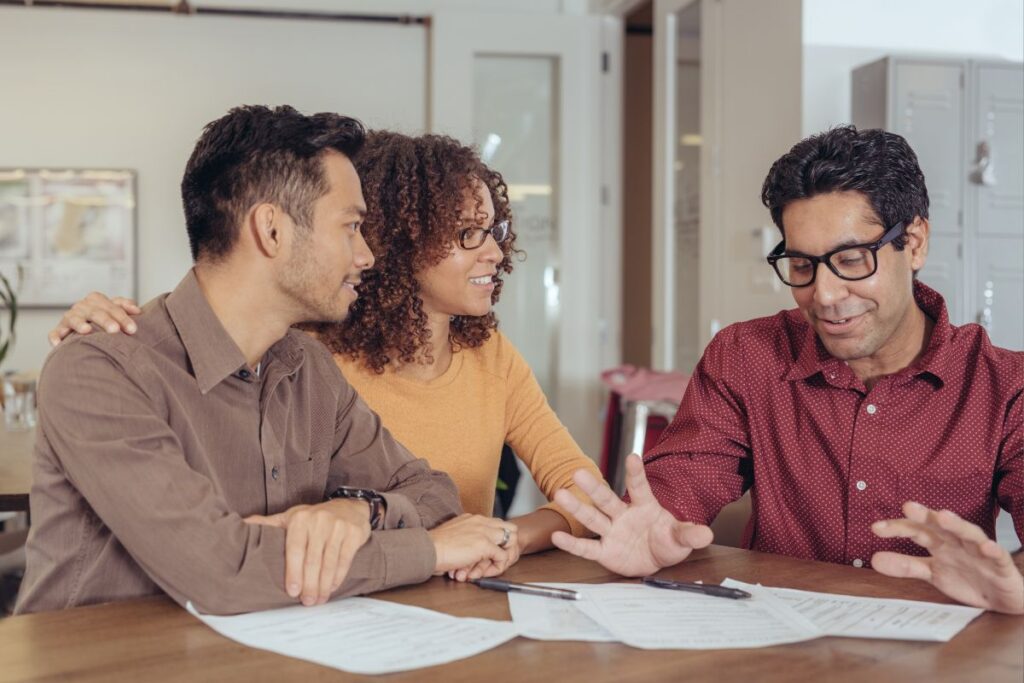 Um casal atento ouvindo um consultor financeiro ou mentor, que gesticula enquanto explica algo sobre documentos na mesa.