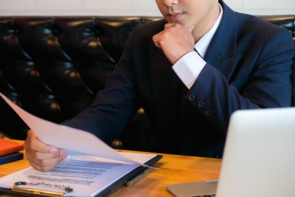 Um homem de terno escuro e camisa branca sentado a uma mesa de madeira, segurando um documento e olhando pensativamente para ele. Um laptop prateado está aberto ao seu lado.