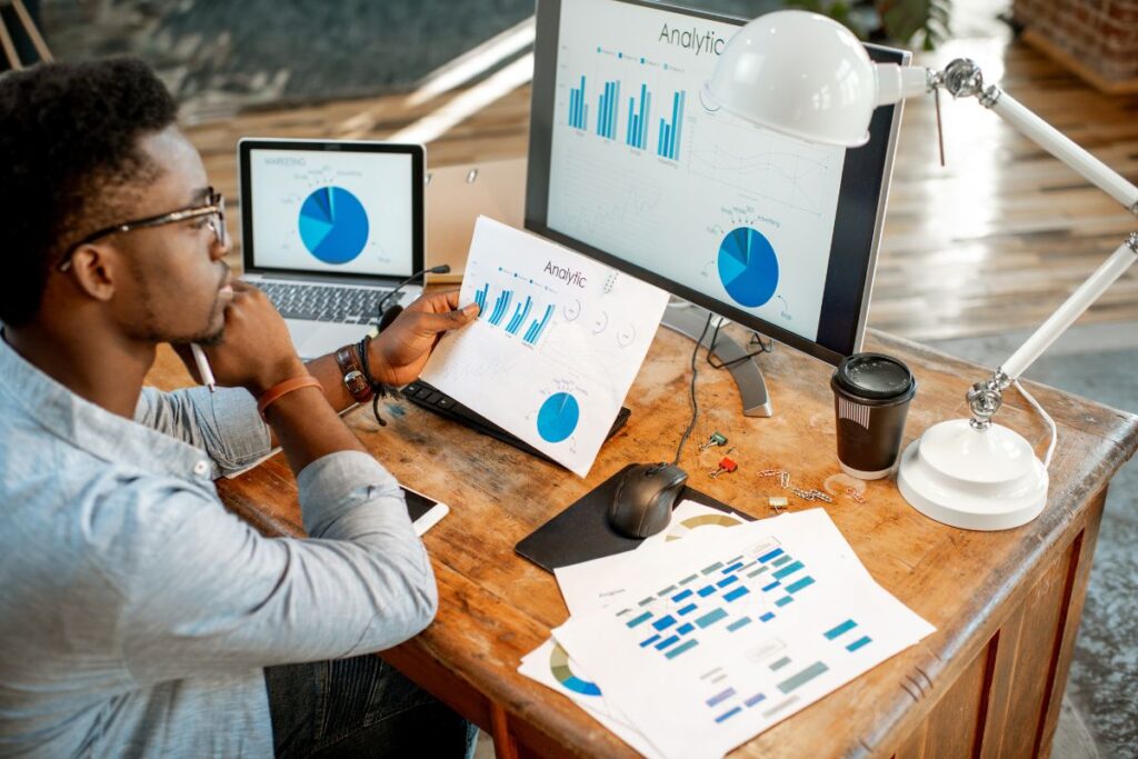 Profissional analisando gráficos e dados de marketing digital em um escritório doméstico com múltiplos monitores.