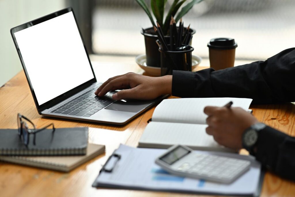 Homem digitando em um laptop com tela em branco e tomando notas em um escritório doméstico.