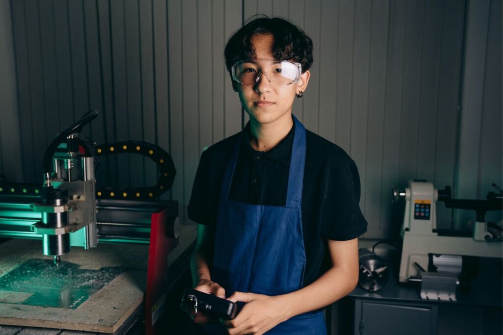 Jovem aprendiz confiante usando equipamentos de proteção individual em um ambiente de oficina industrial com máquinas CNC.