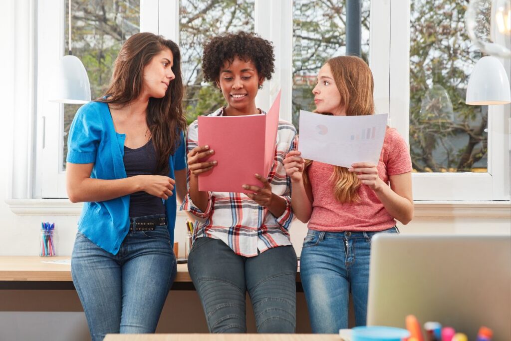 Três jovens aprendizes multirraciais discutem documentos em um ambiente de trabalho moderno e iluminado.