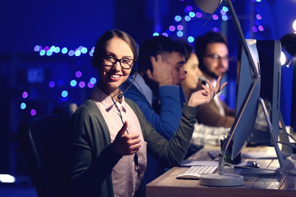 Uma equipe de atendimento ao cliente trabalhando em computadores à noite, com uma mulher em primeiro plano sorrindo e mostrando um sinal de positivo, representando habilidades de comunicação e trabalho em equipe no ambiente de trabalho.