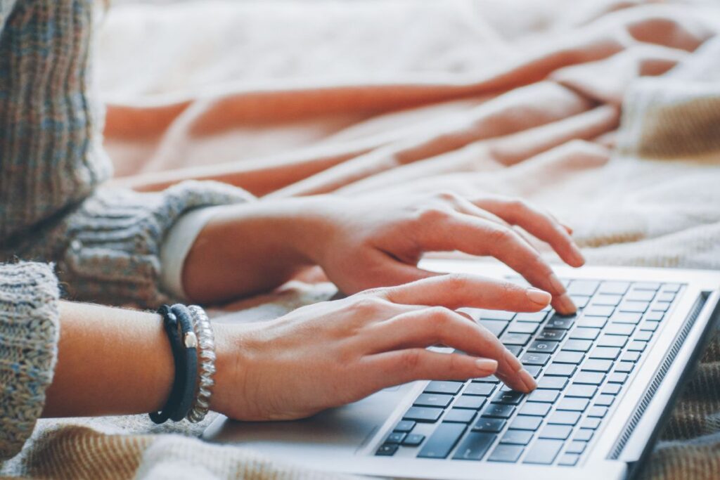 Close-up de mãos femininas digitando em um laptop sobre uma superfície aconchegante, simbolizando o aprendizado e o trabalho online para o desenvolvimento de habilidades.