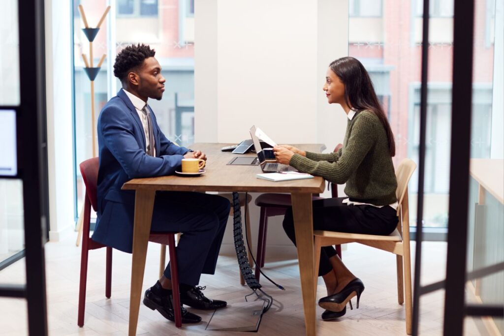 Um homem e uma mulher, ambos profissionais, sentados à mesa um de frente para o outro durante uma entrevista de emprego. O homem, em um terno azul, está sendo entrevistado pela mulher, vestida casualmente elegante, em um ambiente de escritório moderno com luz natural e janelas amplas.