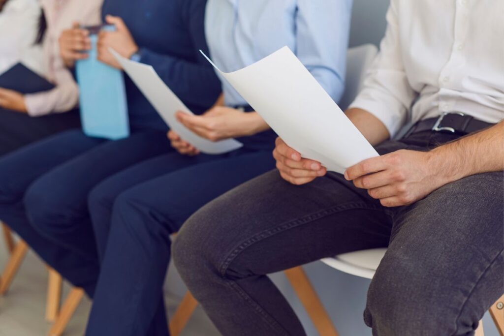 Candidatos aguardam em fila, segurando seus currículos e outros documentos para entrevistas de emprego, sentados em cadeiras de escritório em uma sala de espera com luz natural.