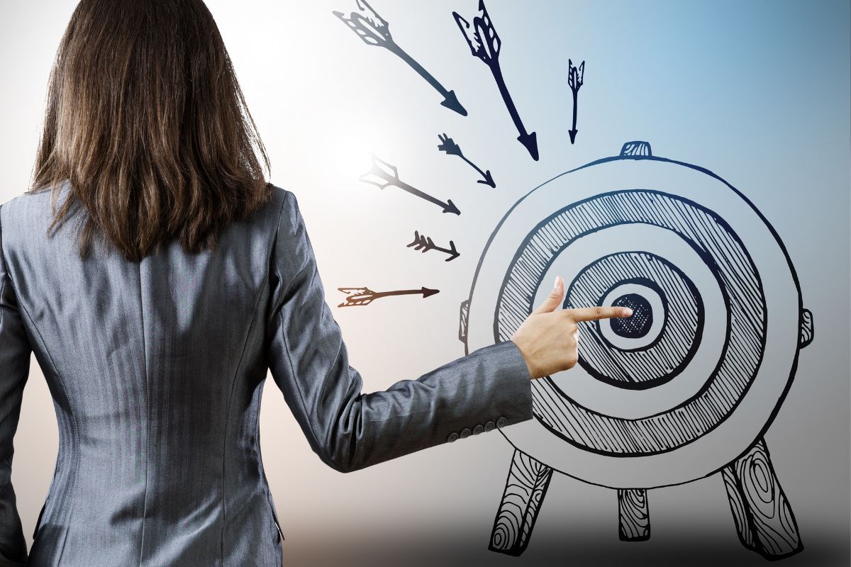 Uma mulher apontando para o centro de um alvo desenhado, com várias flechas dispersas ao redor.