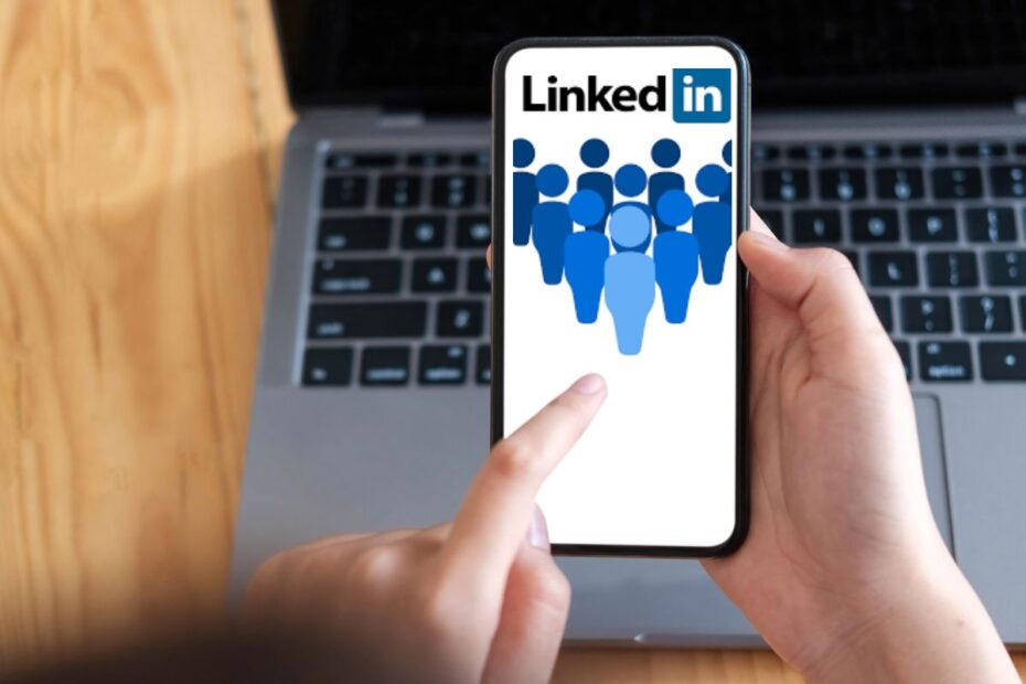 Mãos segurando um smartphone com o aplicativo do LinkedIn aberto, exibindo ícones de pessoas em azul sobre um fundo branco, com um teclado de laptop em segundo plano.