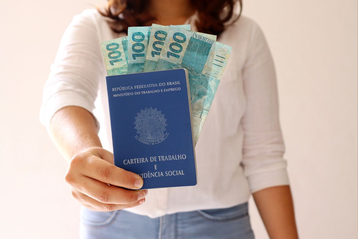 Pessoa segurando uma Carteira de Trabalho e Previdência Social com notas de cem reais saindo dela, representando o 13º salário.