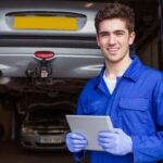 Jovem aprendiz em oficina mecânica sorri, segurando um tablet, com um carro elevado ao fundo.