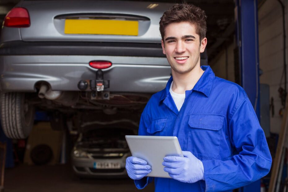 Jovem aprendiz em oficina mecânica sorri, segurando um tablet, com um carro elevado ao fundo.