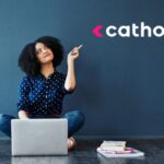 Jovem profissional sorridente apontando para logo da Catho enquanto usa laptop para criar currículo