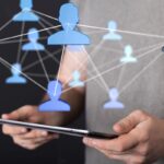 Uma pessoa segurando um tablet, com uma projeção digital de ícones de pessoas azuis conectadas por linhas, ilustrando o uso da tecnologia para networking.