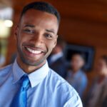 Homem negro, sorridente e bem-humorado, vestindo camisa social azul e gravata, em ambiente corporativo. Representa a satisfação e o bem-estar no trabalho, que as férias podem impulsionar.