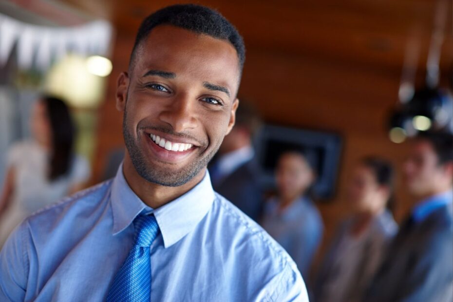 Homem negro, sorridente e bem-humorado, vestindo camisa social azul e gravata, em ambiente corporativo. Representa a satisfação e o bem-estar no trabalho, que as férias podem impulsionar.