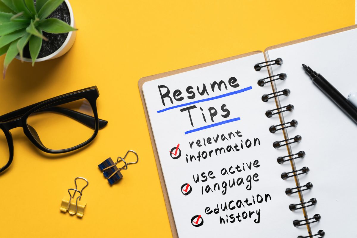 Uma caneta preta e um caderno aberto com "Resume Tips" (Dicas de Currículo) escrito na página e bullet points sobre informações relevantes, linguagem ativa e histórico educacional. Há também óculos escuros e clipes de papel sobre uma superfície amarela.