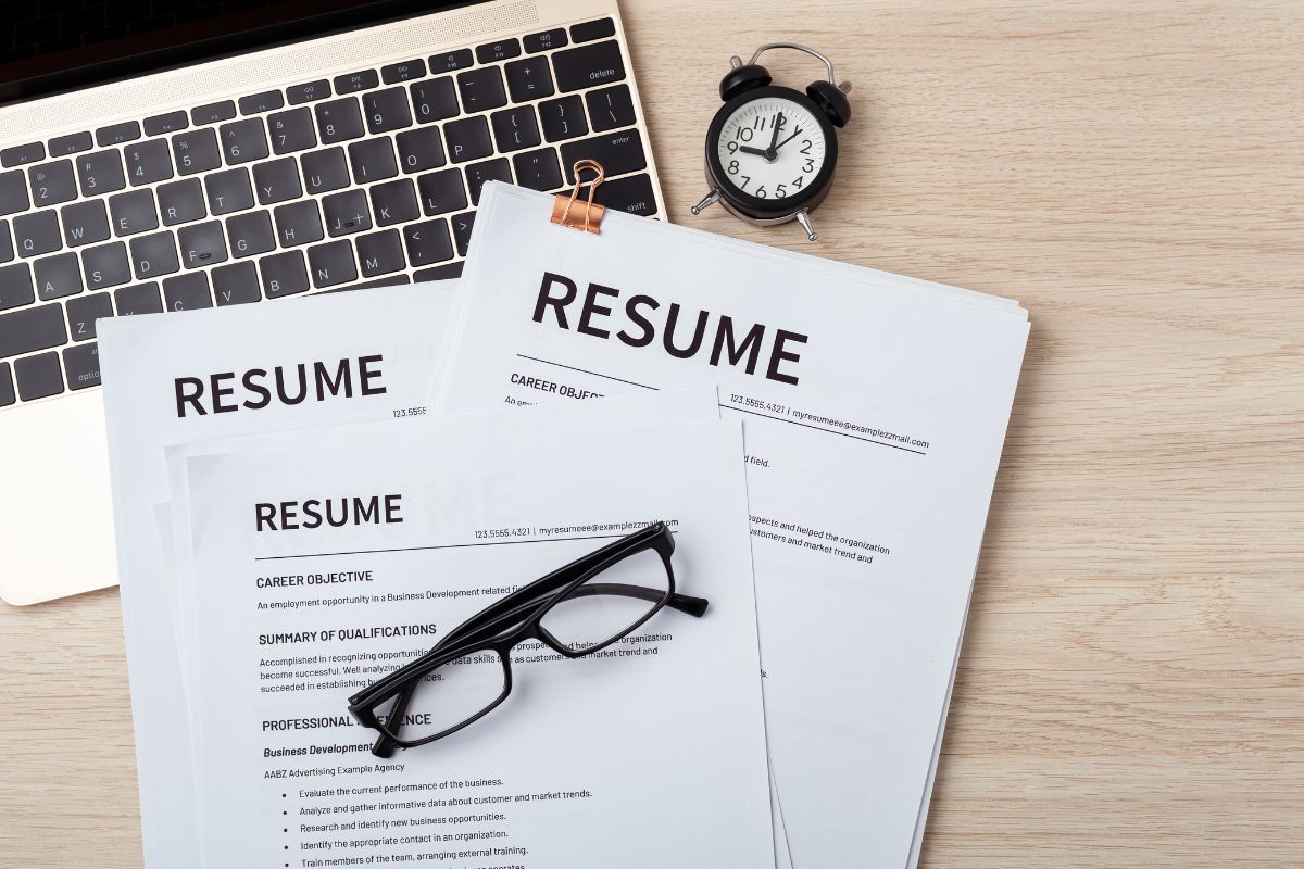 Pilha de currículos impressos sobre uma mesa de madeira, ao lado de um laptop, óculos e um despertador.